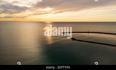 Drohnenblick auf die Morgensonne, die hinter Wolken über einer ruhigen Meeresbucht mit Fischfarmen im Hintergrund aufgeht Stockfoto