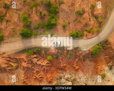 Aus der Vogelperspektive eines Einzelwagens navigiert eine kurvige Feldstraße durch die trockenen, rotfarbenen Landschaften, die einen starken Kontrast zur sporadischen grünen Vegetation bilden. Stockfoto