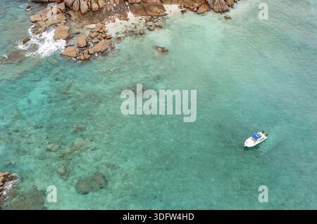 Aus der Vogelperspektive sehen Sie das durchscheinende türkisfarbene Wasser entlang der felsigen Küste, wo ein einsames Boot ruhig schwimmt, die Seychellen. Stockfoto