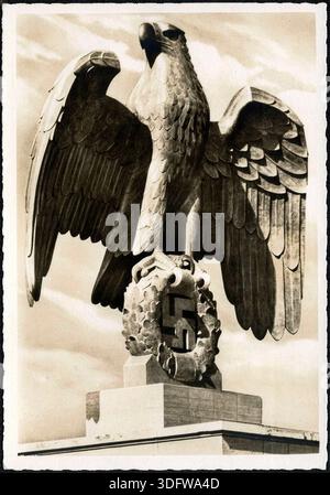 Diese Vintage-Fotografie aus den 1930er bis 1940er Jahren zeigt eine monumentale Steinskulptur eines Adlers, der auf einem verkranzten Hakenkreuz-Symbol thront. Die imposante Statue, wahrscheinlich ein Nazi-deutsches Emblem, steht vor einem bewölkten Himmel und erinnert an die düstere Atmosphäre der Epoche. Das Bild scheint von einem historischen Dokument oder einer Postkarte zu stammen. Stockfoto