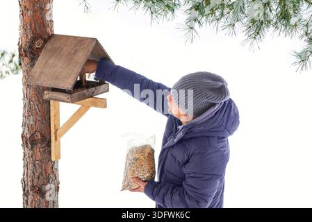 Mann mittleren Alters füllen Holzvogelfutter mit Samen auf Kiefer im Winterpark auf. Fütterung von Vögeln und Eichhörnchen im kalten Winter. Aktive Pensionierung und Unterstützung für Wildtiere. || Modell freigegeben Stockfoto
