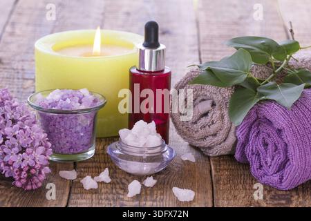 Lila Blüten, Schalen mit Meersalz, brennende Kerze, rote Flasche mit Aromaöl und Handtücher für Bad Verfahren mit grünen Blättern auf Holzplatten Stockfoto
