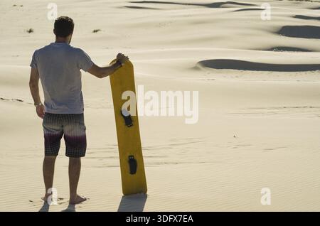 Ein junger Mann auf dem Rücken, der auf die Sanddünen blickt und sich auf das Sandboarden vorbereitet Stockfoto
