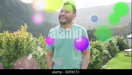 Stehender Mann im mintfarbenen Polo, lächelnd und blickend nach oben im Garten mit Hecke, Berg, Linsenfleck Stockfoto