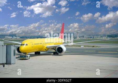 Ein hellgelbes Flugzeug mit rotem Schwanz wartet auf der Landebahn an einem stark frequentierten Flughafen. Die Bodencrew bereitet sich auf den Start vor, während die Sonne in einem klaren Licht untergeht Stockfoto