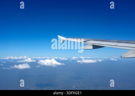 Ein Flugzeugflügel erstreckt sich in einen hellblauen Himmel voller weicher weißer Wolken und fängt die heitere Schönheit der Flugreisen an einem sonnigen Tag ein. Stockfoto
