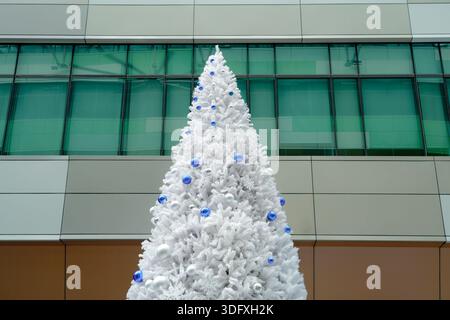 Vor einem modernen Gebäude steht ein hoher weißer Weihnachtsbaum, der mit blauen Ornamenten und funkelnden Lichtern geschmückt ist und eine festliche Atmosphäre schafft Stockfoto