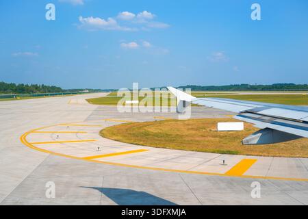 Der Blick von einem Flugzeugfenster zeigt den Flügel und eine Start- und Landebahn des Flughafens an einem sonnigen Tag. Gras und Bäume sind in der Ferne unter der hellblauen Ske zu sehen Stockfoto