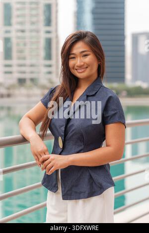 Eine asiatische Frau steht selbstbewusst auf einem Geländer in einer intelligenten Stadt und zeigt ihren professionellen Stil, während sie in einer geschäftigen Umgebung im Freien arbeitet. Stockfoto