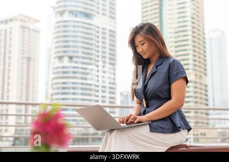 Asiatische Frau konzentriert sich auf ihren Laptop, während sie draußen in einer geschäftigen Stadt mit hohen Gebäuden in der Nähe sitzt. Sie engagiert sich für ihre Arbeit. Stockfoto