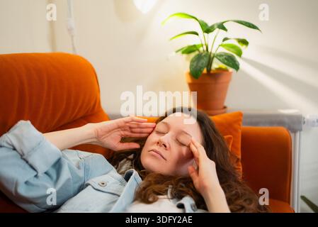 Frau, die die Augen schließt und die Hände auf den Schläfen drückt, intensive Kopfschmerzen, Schmerzen und Erschöpfung durch Stress oder Krankheit spürt, Linderung sucht, während sie sich auf einem bequemen Sofa zu Hause zurücklehnt Stockfoto