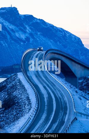 Ein Auto, das über eine gekrümmte Brücke auf einer vereisten Winterstraße fährt, während der blauen Stunde Berge im Hintergrund aufsteigen. Stockfoto