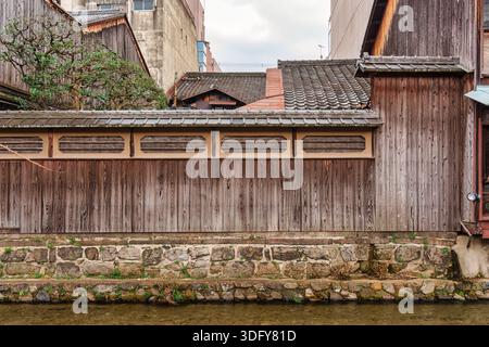 Äußere eines alten traditionellen japanischen Holzhauses mit Fliesendach, Steinmauer und Baum am Kanal in Kyoto, Japan Stockfoto