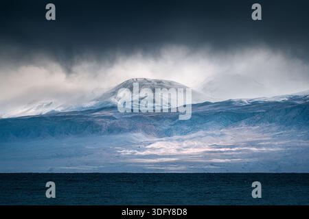 Dramatisch stimmungsvolle Landschaft mit schneebedeckten Bergen, Wildnis und bewölktem Himmel über dem dunklen Ozean in stürmischem Klima Stockfoto