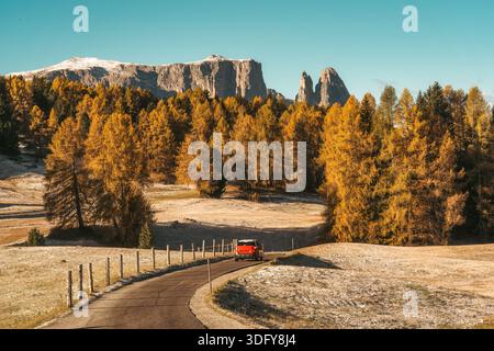 Rotes Auto, das im Herbst auf der Seiser Alm in Italien auf einer kurvenreichen Straße durch goldenen Lärchenwald und Dolomiten fährt Stockfoto