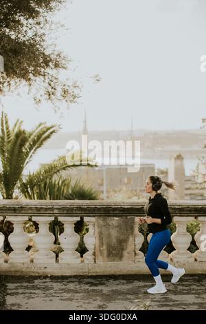 Eine Frau joggt an der Küste in Malta. Sie trägt Trainingskleidung und Kopfhörer und läuft auf einem Weg mit Blick auf das Wasser und die Gebäude. Das ist es Stockfoto
