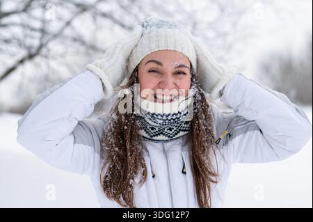 Porträt einer fröhlichen Frau, die ihre Strickmütze in einem verschneiten Winterpark anpasst Stockfoto