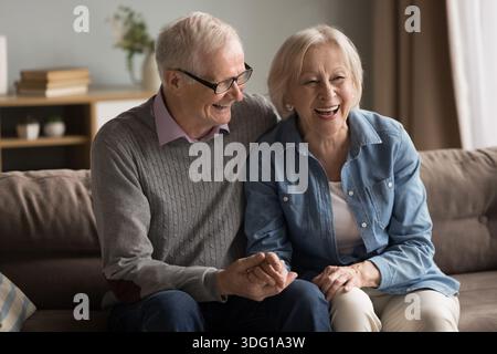 Romantisches, fröhliches, liebevolles Ehepaar im Alter, das sich auf ein Sofa kuschelt und die Hände hält Stockfoto