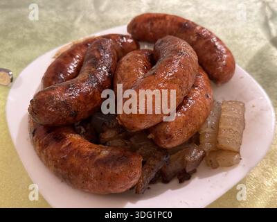 Saftige, braune Würstchen auf einem weißen Teller, ergänzt durch karamelisierte Zwiebeln; ein köstliches, herzhaftes Gericht mit reichen Aromen, das eine Versuchtin präsentiert Stockfoto