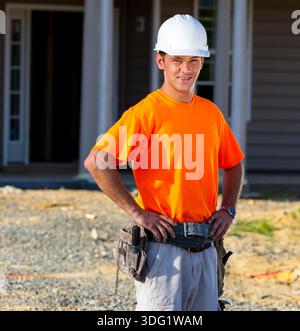 Auf einer sonnendurchfluteten Baustelle steht ein leitender Bauarbeiter vor einem aufstrebenden Familienhaus und studiert mit Fokus und Selbstvertrauen Baupläne, um jeden Fall sicherzustellen Stockfoto