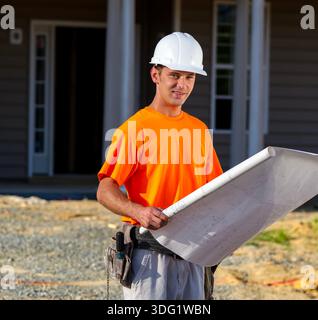 Auf einer sonnendurchfluteten Baustelle steht ein leitender Bauarbeiter vor einem aufstrebenden Familienhaus und studiert mit Fokus und Selbstvertrauen Baupläne, um jeden Fall sicherzustellen Stockfoto