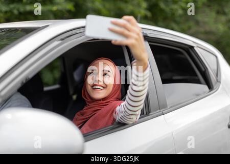 Glückliche junge arabische Dame in Hijab nimmt Selfie am Telefon im offenen Fenster im Auto, Ehemann fährt, im Freien Stockfoto