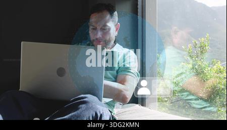 Arbeitender Mann im grünen Polo, der auf silbernem Laptop am Fenster mit digitaler Kugel schreibt, Becher Stockfoto