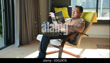 Lesender reifer Mann, der sich in einem hölzernen Loungesessel neben Glastüren liegend, Buch in der Hand hielt, Brille trug Stockfoto