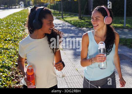 Zwei verschiedene Frauen in Activewear und Kopfhörern spazieren und plaudern auf einem sonnigen Parkweg, lächeln, halten sich mit einer Wasserflasche versorgt und genießen dabei die Passform Stockfoto