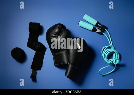 Ein Paar schwarze Boxhandschuhe mit Armbändern und Sprungseil auf blauem Hintergrund Stockfoto