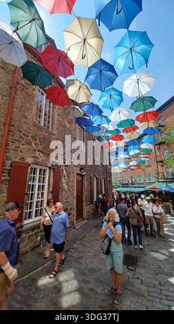 Montreal, QC, Kanada - 7. August 2025: Leuchtende Regenschirme schaffen eine farbenfrohe Decke über einer historischen Kopfsteinpflasterstraße, bei der die Menschen die Sonne genießen Stockfoto