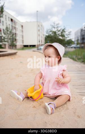 Entzückendes Kleinkind in rosa Kleid mit Sonnenhut, das fröhlich auf dem urbanen Sandspielplatz neben Wohngebäuden unter teilweise bewölktem Himmel spielt Stockfoto
