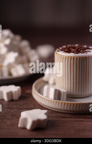 Keramikbecher voller cremiger heißer Schokolade, gekrönt mit Schokoladenspänen und umgeben von sternförmigen Marshmallows auf einem rustikalen Holztisch, coa Stockfoto