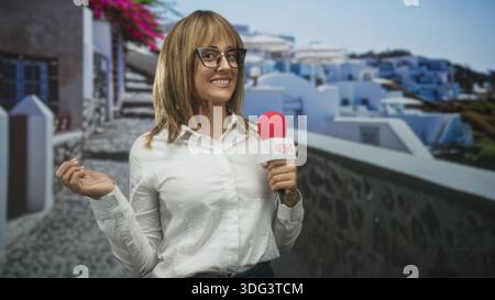 Reporterin mit rotem Mikrofon und Nachrichtenfahne, lächelnd und gestisch mit erhobener Hand, mit Brille und weißem Hemd am Studio-Set; vertrauensvoll Stockfoto