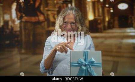 Die Seniorin zeigt mit dem Finger auf eine verpackte Geschenkbox mit blauem Band im prunkvollen Gebäude; Freude am Feiern. Stockfoto