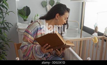 Junge hispanische Frau mit Brille sitzt auf einem Stuhl und liest ein Buch über einer Babykrippe in der Nähe von Pflanzen und einem hängenden Mobile im Studio; ruhige Verbindung Stockfoto