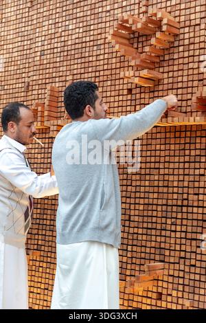 Al-Balad, Dschiddah, Saudi-Arabien - 23. Dezember 2025: Zwei Männer arbeiten zusammen, um Holzblöcke auf einem großen interaktiven Holzkunstinstallat zu platzieren und auszurichten Stockfoto