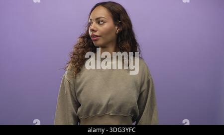 Das Gesicht der jungen Frau sieht richtig aus und trägt ein lässiges, verkürztes Sweatshirt und einen kleinen Ohrring mit Nieten im lila Studio; ruhiger Zuversicht-Optimismus. Stockfoto