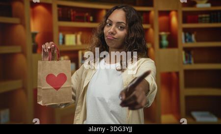 Die junge afroamerikanische Frau hält eine Papiertüte mit rotem Herzen und zeigt mit dem Finger in das Gebäude; selbstbewusster Trotz. Stockfoto