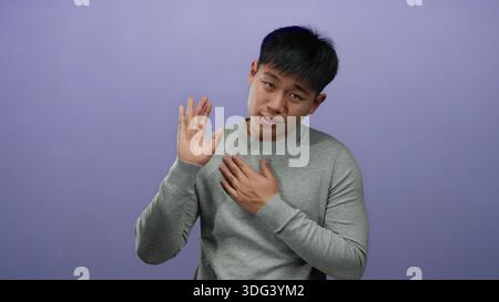 Junger chinesischer Mann im grauen Pullover Gesten aufrichtig mit Hand auf Herz über lila Hintergrund Wand, die Ehrlichkeit und Vertrauen vermittelt. Stockfoto