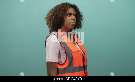 Junge Technikerin mit orangefarbener reflektierender Sicherheitsweste, die im Studio mit blauem Hintergrund steht und blickt; Selbstvertrauen. Stockfoto