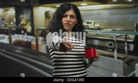 Junge brünette Frau zeigt mit dem Finger nach vorne, während sie im Küchengebäude einen roten Becher hält; Missbilligung. Stockfoto