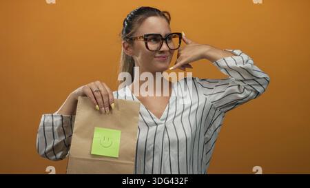 Frau, die eine Brille trägt, eine Papiertüte mit einem lächelnden Haftnotiz hält und mit dem Finger ins Studio zeigt; freundliches Vertrauen. Stockfoto