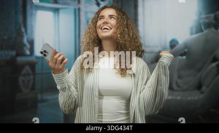 Junge Frau, die das Smartphone mit erhobener Hand und breitem Lächeln im Foyer hält, Hand sichtbar und lockiges Haar; freudiges Vertrauen. Stockfoto