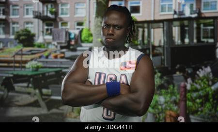 Die Arme des Mannes kreuzten sich, tragend ein us-Basketballtrikot und blaues Armband, Flaggenaufnäher sichtbar, stehend in einem städtischen Straßencafé mit Bänken und Pflanzgefäßen; def Stockfoto