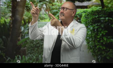 Mann Doktor, Senior hispanic im Laborkitt zeigt mit beiden Händen den Finger nach oben in die Nähe des Gartenturms am Gebäude, trägt Brille und Taschenstifte; Kuriosit Stockfoto