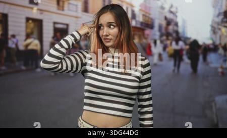 Die Frau zeigt den Finger mit besorgtem Ausdruck auf den Tempel, trägt ein gestreiftes Hemd und einen Nietengürtel inmitten einer überfüllten Stadtstraße; Zweifel an Verwirrung. Stockfoto