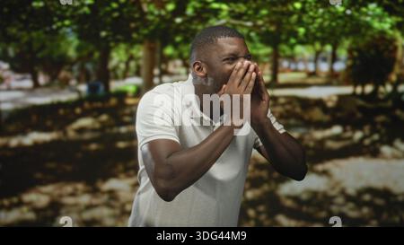 Junger schwarzer Mann, der die Hände an Nase und Mund drückt, während er auf einem schattigen Waldweg unter Bäumen niest; Allergiebeschwerden. Stockfoto