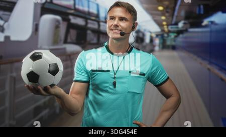 Schiedsrichter hält Fußball in der Hand, Pfeife sichtbar und Headset-Mikrofon im Gebäudekorridor; Vertrauensmacht. Stockfoto