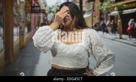 Frau mit Hand auf der Stirn für Kopfschmerzen auf der Straße, mit weißer, bestickter Bluse, zuwinkenden und reibenden Schläfen; Stress-Müdigkeit-Frustration. Stockfoto